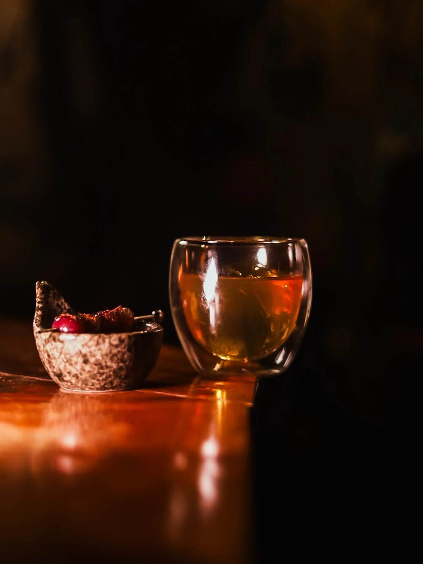 Amber cocktail in a double-walled glass beside a small bowl of garnishes on the warmly lit bar.
