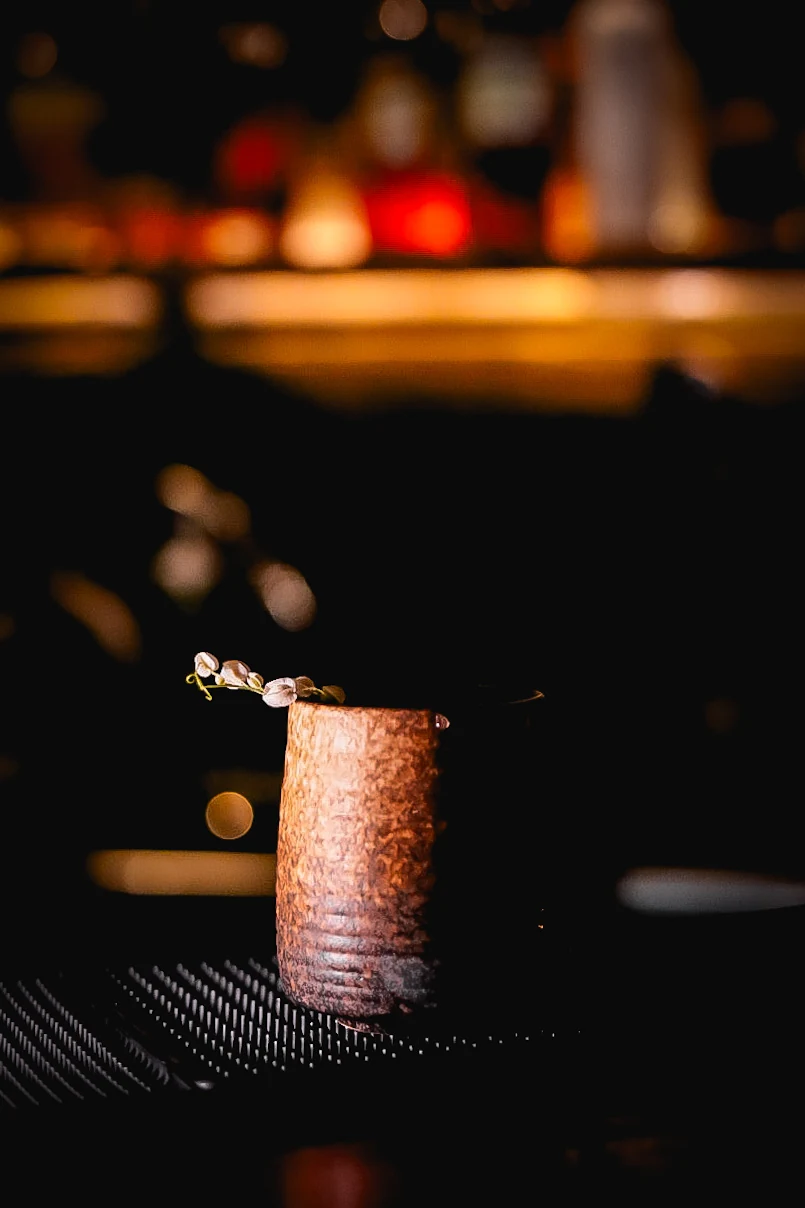 Cocktail in a textured copper-colored cup with a pale floral garnish against warm bar lights.