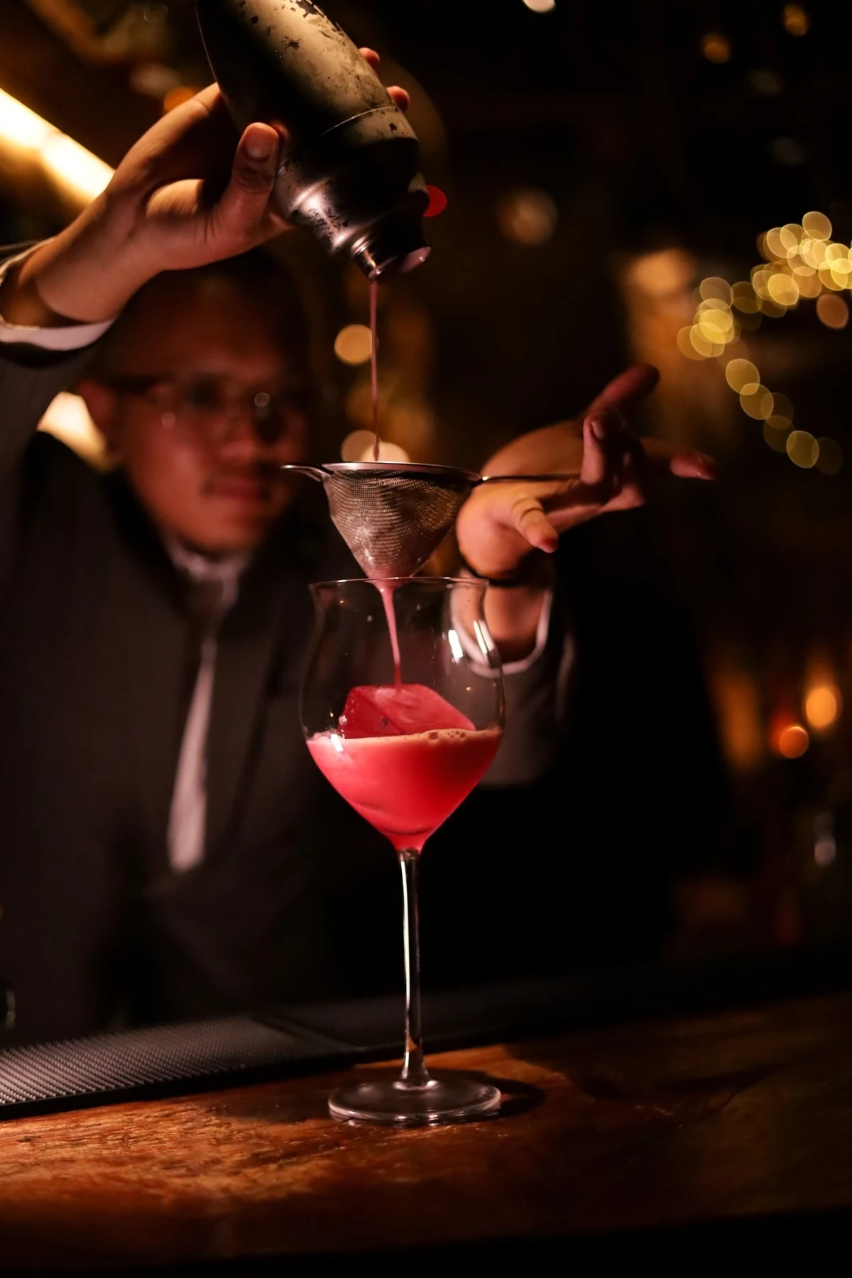 A bartender pouring a red cocktail through a fine strainer against warm golden lights.