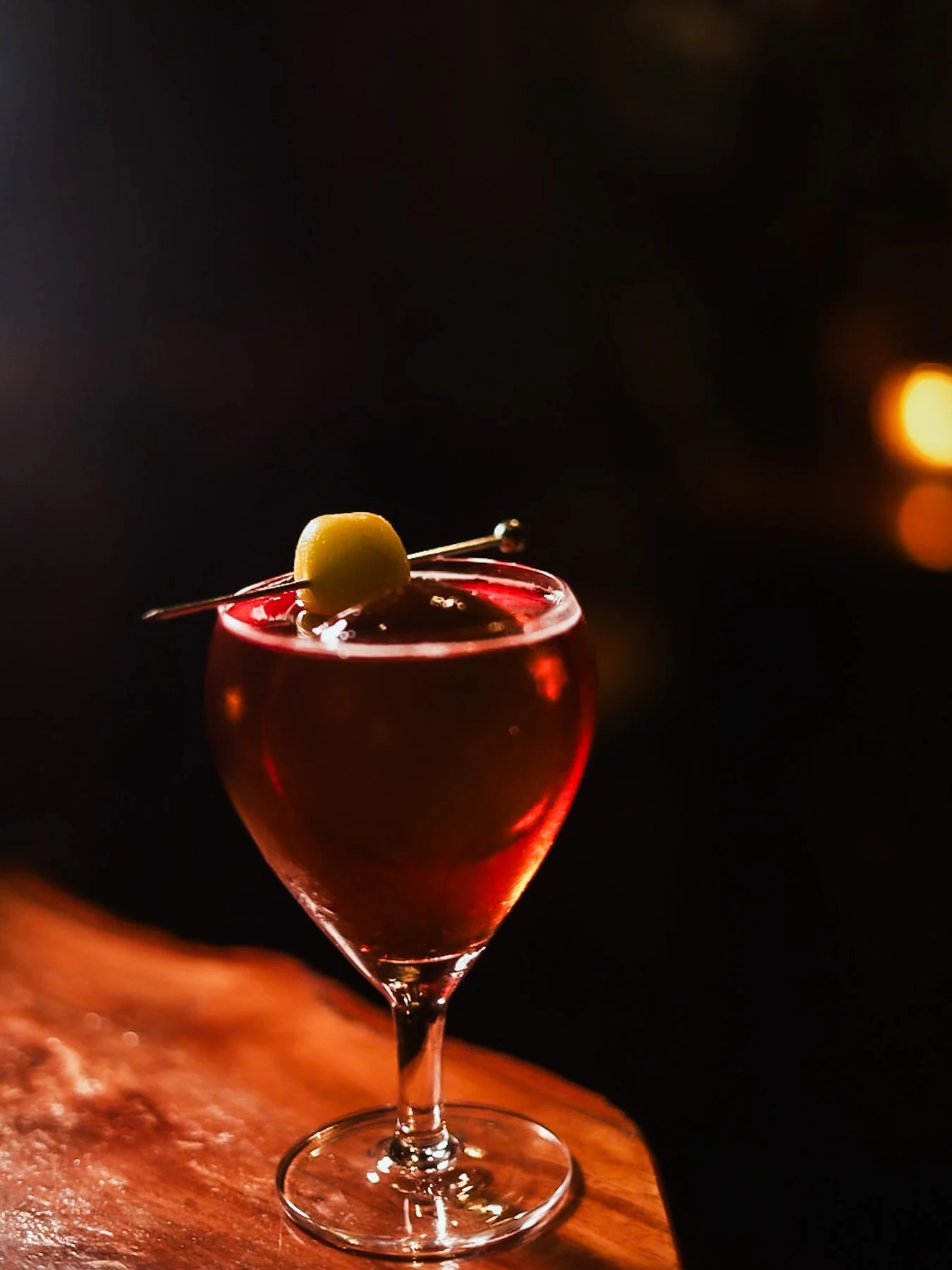 Deep red cocktail in a stemmed glass with a single olive garnish on the wooden bar.
