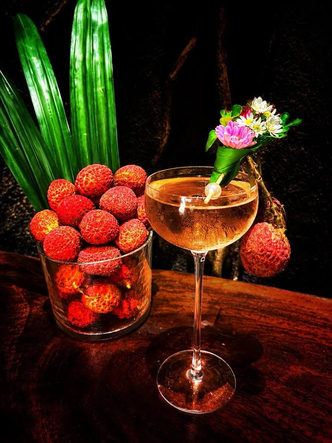 Amber cocktail with a floral garnish beside fresh lychees.