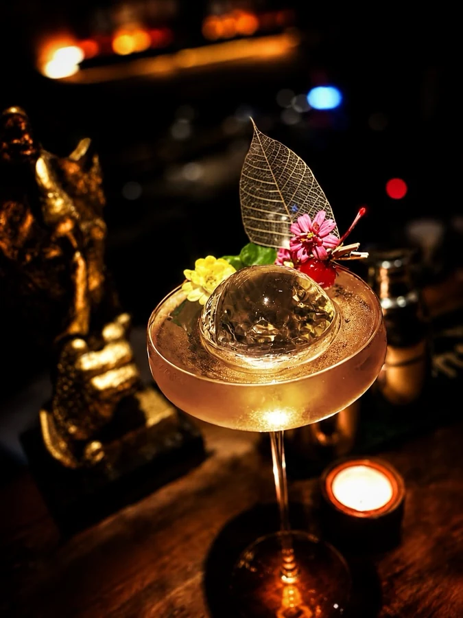 Golden cocktail topped with a clear bubble, flowers, and a delicate leaf.