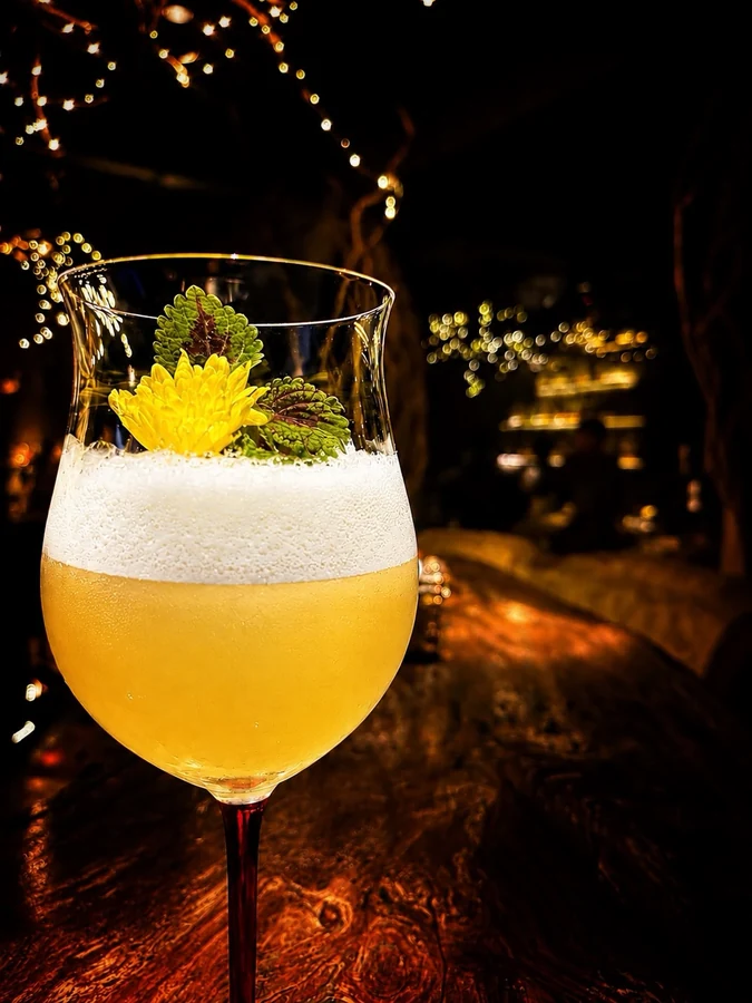 Golden cocktail with foam and a yellow flower garnish.