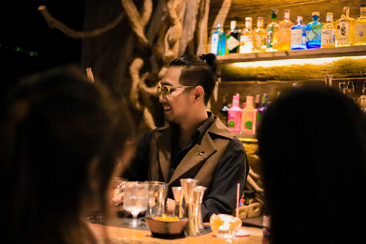 Smiling bartender behind the wooden counter beneath warm bottle shelves and twisting roots.