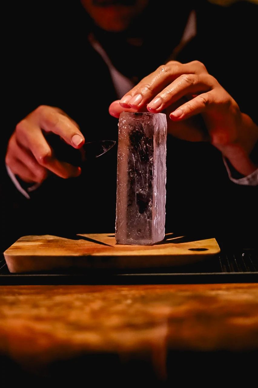 Bartender hand-carving a tall clear ice block on a wooden board.