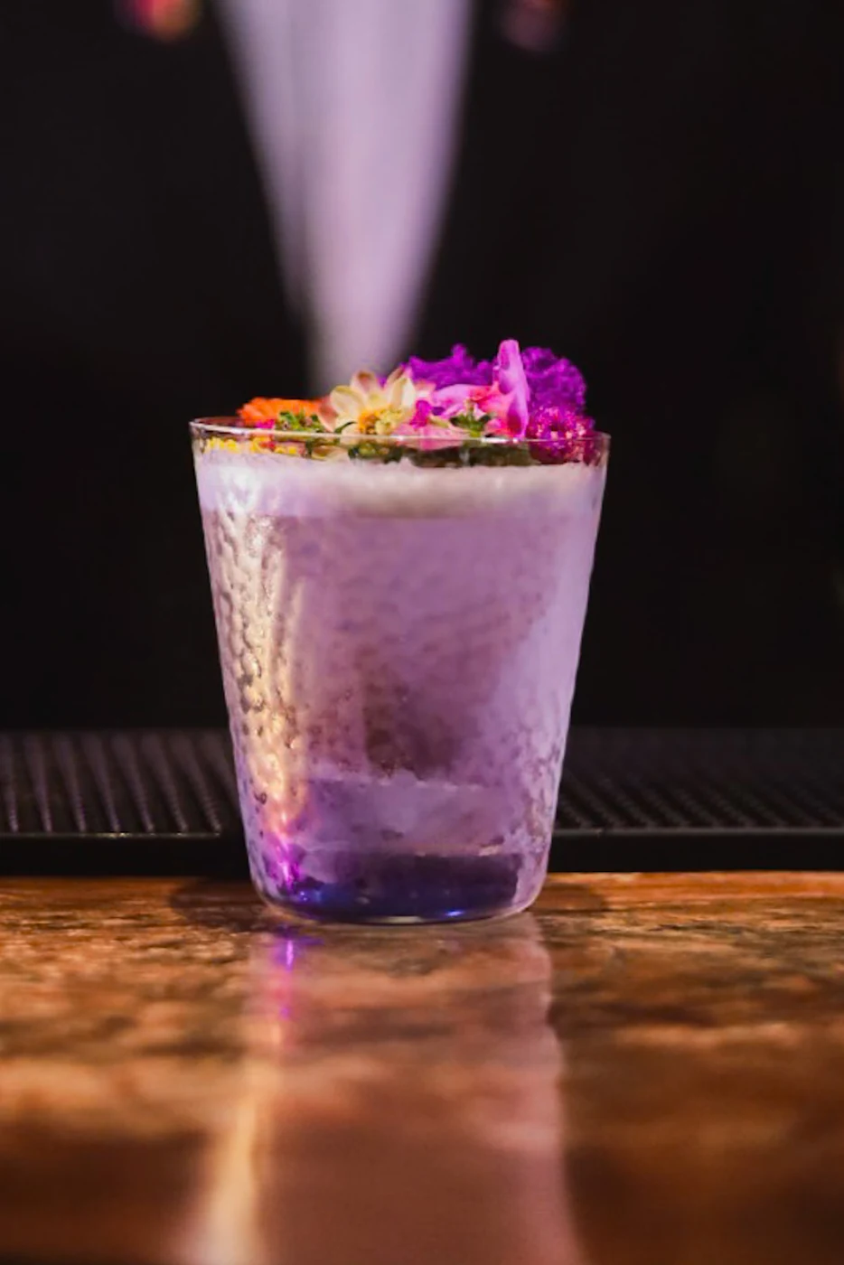 Purple cocktail with a white foam cap and colorful flower garnish on the wooden bar.
